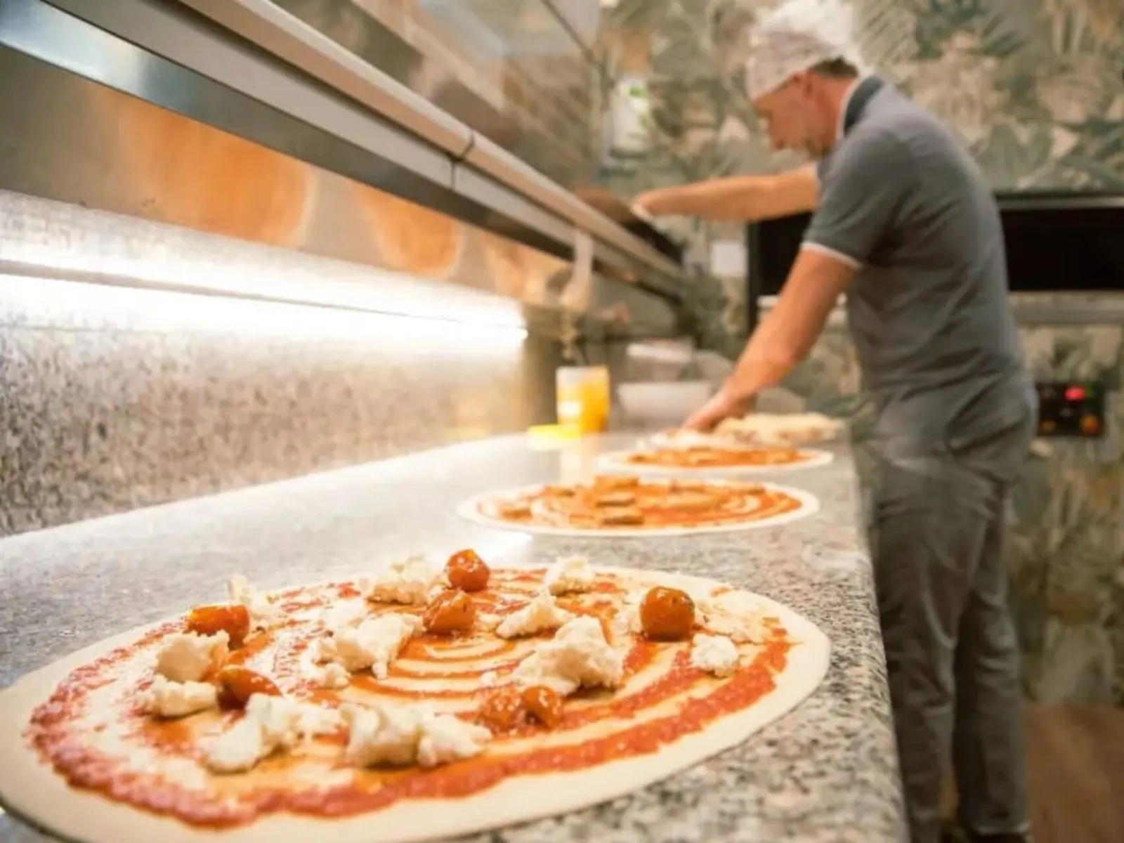 Pizze appena stese sul bancone, con pomodoro, bufala e pomodorini; dietro, il pizzaiolo al lavoro