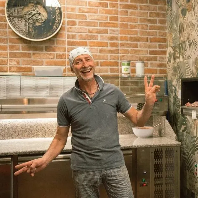 Il pizzaiolo sorridente al banco, davanti al muro di mattoni